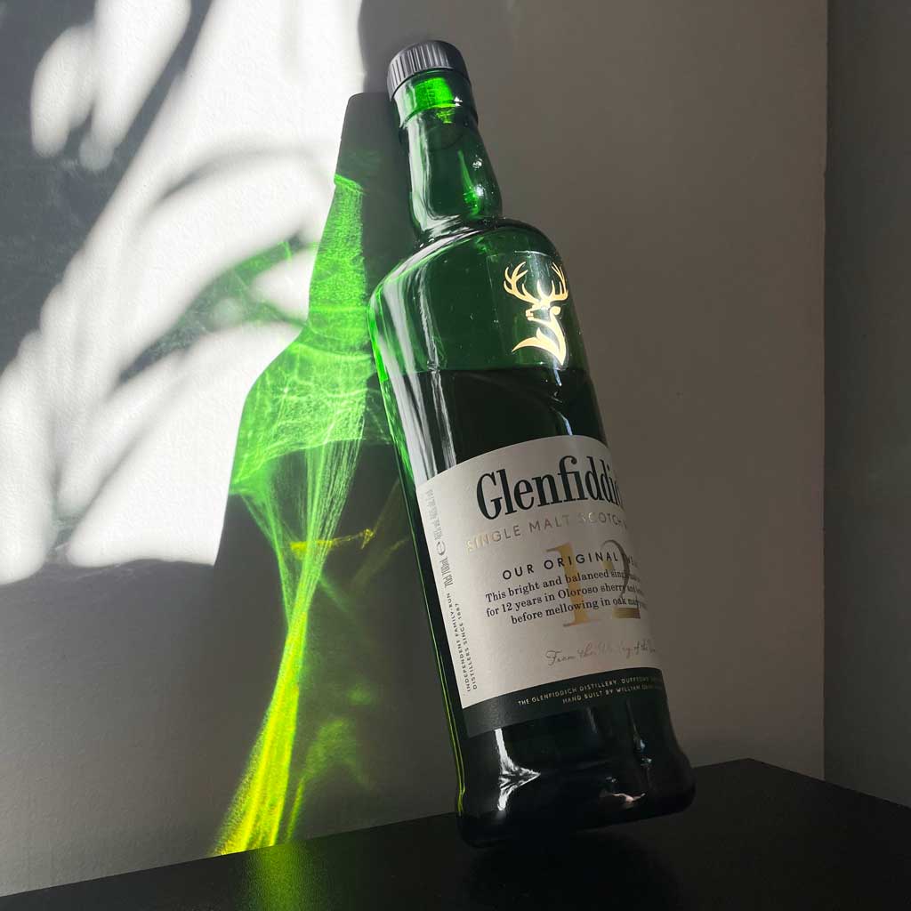 Bottle of 12 year old Glenfiddich whisky leaning against white wall in bright sunlight
