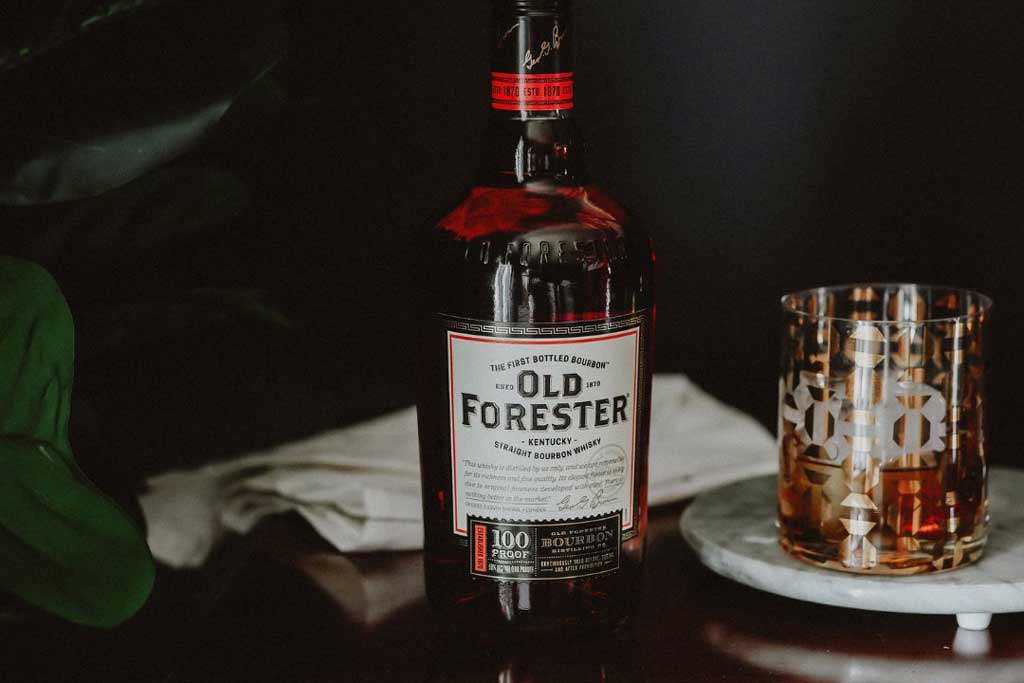 Bottle of Old Forester 100 proof bourbon on drinks cabinet beside rocks glass and green plant