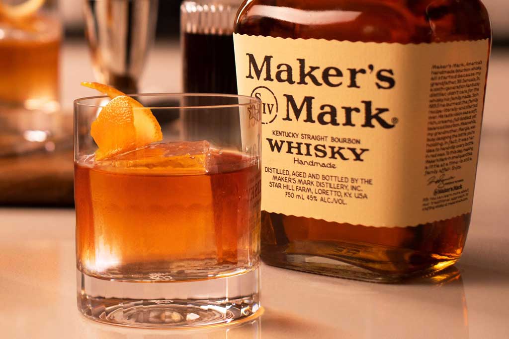 Close view of an Old Fashioned cocktail beside a bottle of Makers Mark bourbon