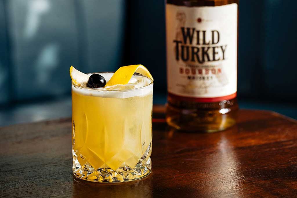 Golden yellow cocktail in rocks glass beside Wild Turkey 101 whiskey bottle