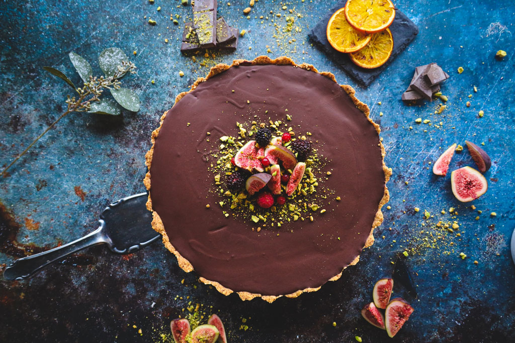 Aerial view of chocolate tart surrounded by fresh figs and oranges on blue table surface