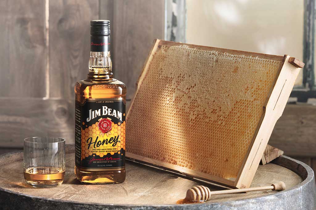 Bottle of Jim Beam honey on top of whiskey barrel beside honeycomb and drinking glass