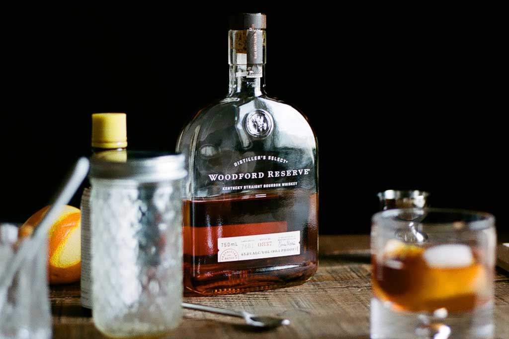 Bottle of Woodford Reserve bourbon on wooden table beside cocktail utensils and drinking glass