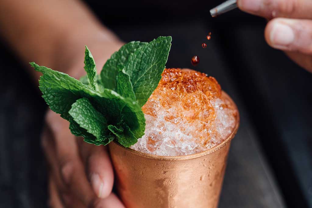 Person pouring cocktail into copper cup with crushed ice and mint sprig