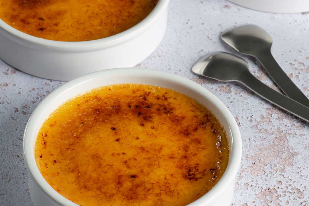 Two crème brûlée desserts in white bowls beside spoons on white table