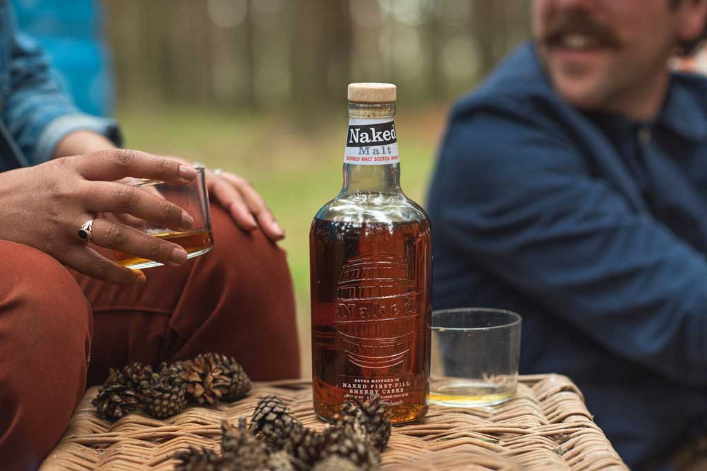 Two men sitting beside bottle of Naked Malt whisky on top of hamper outside