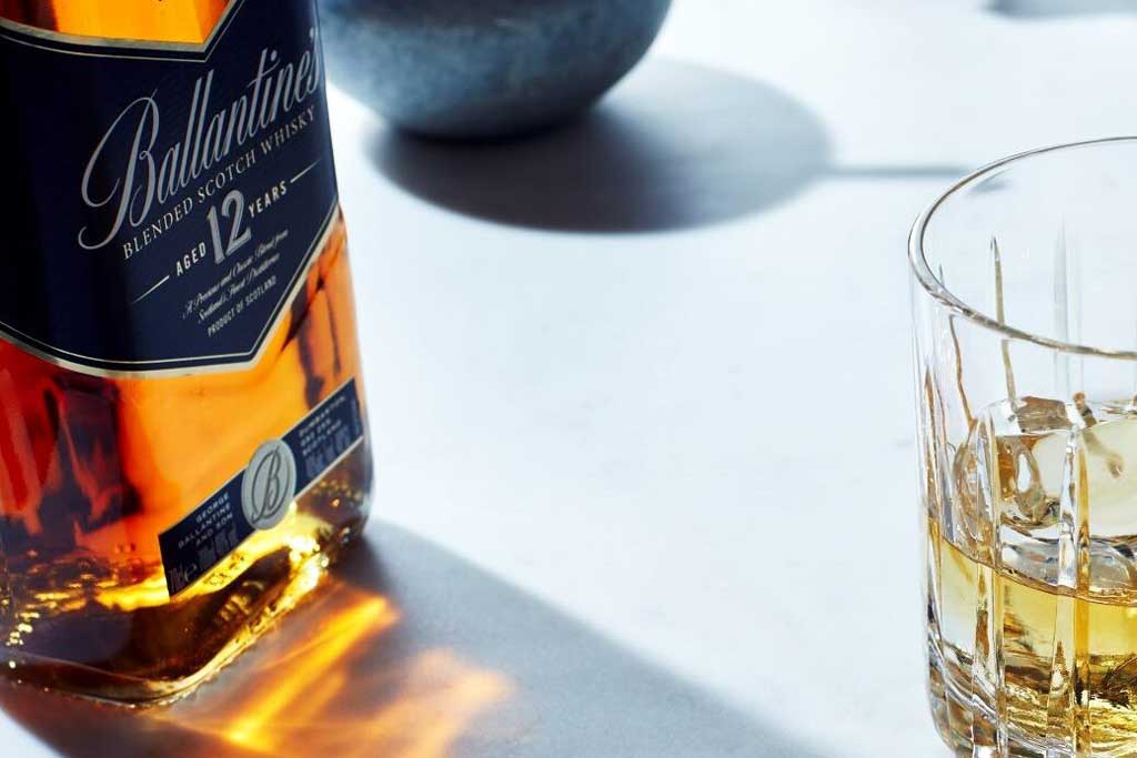 Bottle of Ballantine's 12 year old blended Scotch whisky on white table beside rocks drinking glass
