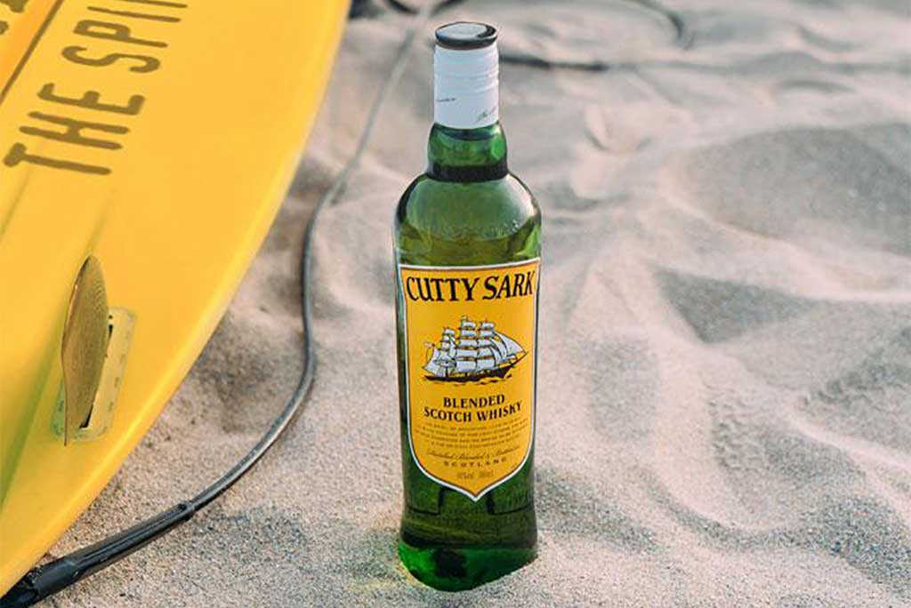 Bottle of Cutty Sark blended Scotch whisky on sandy beach beside yellow surf board